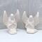 Set of 2 paintable ceramic angel statues sitting on a light blue table with an ecru lacy curtain behind them.